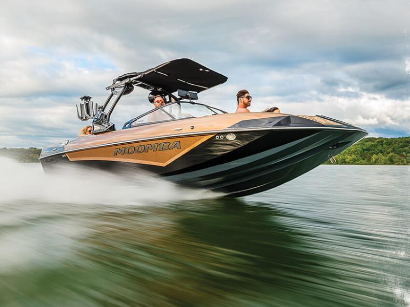 Two men riding a brown & black 2020 Moomba Kaiyen wakeboard boat through the water on a partly cloudy day.