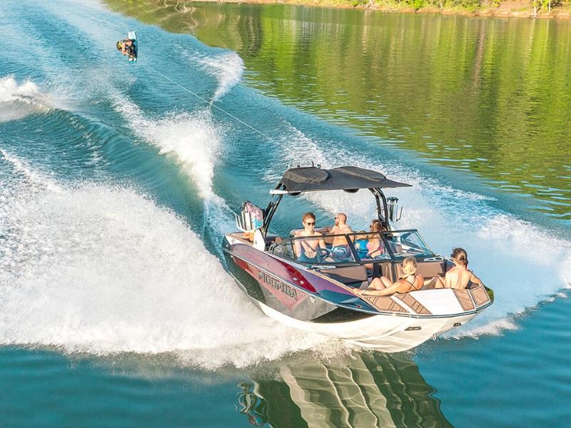 Five people in a red 2021 Moomba Makai wakeboard boat riding in a lake as a person wakeboards behind them.